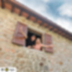 Dave & Jenny Marrs smiling and waving from Campo Sasso's window with wooden shutters. Blue sky above, creating a joyful and welcoming scene.
